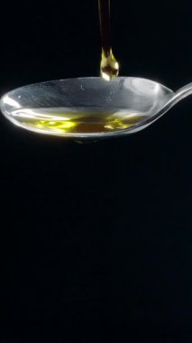 Parlak altın zeytinyağı bir kaşığa dökülüyor ve siyah bir arka planda taşıyor. Yavaş çekim dikey şablonu.
