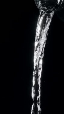 Saf su, siyah arka plandaki cam bir şişeden dökülüyor. Şeffaf su akışı. Yavaş çekim dikey şablonu.