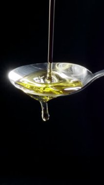 Parlak altın zeytinyağı bir kaşığa dökülüyor ve siyah bir arka planda taşıyor. Yavaş çekim dikey şablonu.