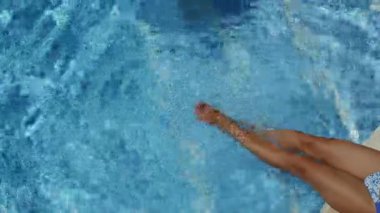 Kadın havuzun kenarında oturuyor. Bacakları suda sallanıyor ve suyu sallıyor. Dalgalar ve küçük dalgalar yaratıyor. Yaz arkaplanı. 