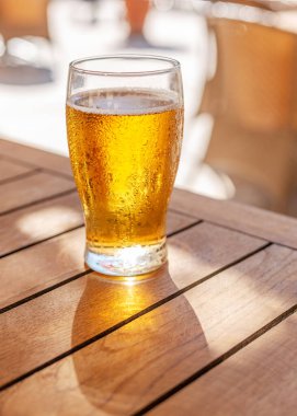 Ahşap masada bir bardak soğuk bira ve arka planda bulanık yaz terası..