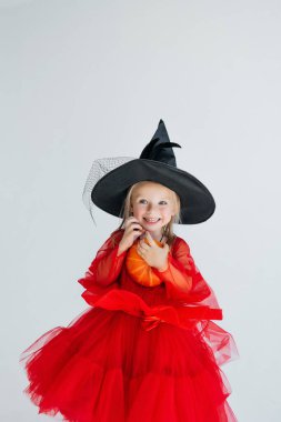 Sivri uçlu şapkalı küçük bir cadı balkabağı tutuyor. Cadılar Bayramı kostümlü bir çocuk mutlu bir şekilde gülümsüyor. Çocuk tatili. beyaz izole arkaplan.