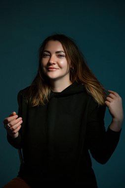 Klasik koyu renk stüdyo portresi. Siyah elbiseli, sandalyede oturan kahverengi saçlı genç bir kadın..