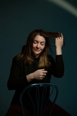 Klasik koyu renk stüdyo portresi. Siyah elbiseli, sandalyede oturan kahverengi saçlı genç bir kadın..