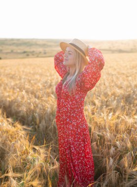 Kafkasyalı sarışın kadın, kırmızı elbiseli ve şapkalı gün batımında altın buğday tarlasında