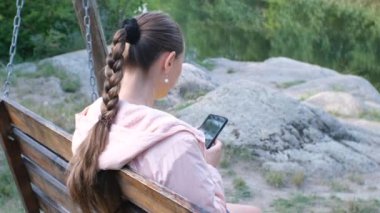 Neşeli orta yaşlı beyaz kadın elinde akıllı bir telefonla bir çadırın yanındaki salıncakta dinleniyor.