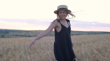 Siyah elbiseli ve şapkalı mutlu orta yaşlı kadın gün batımında buğday tarlasında koşuyor.