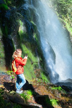 Kırmızı ceketli yürüyüşçü kadın ormandaki bir şelalenin altında fotoğraf çekiyor.