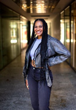 Afro örgülü, gülümseyen siyah bir kadın, arka planda ışık olan bir koridorda kalıyor.