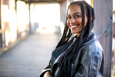 Afro örgülü ve güzel hatları olan genç siyah kadın arka planda bir ışıkla ahşap bir köprüde kalıyor. 