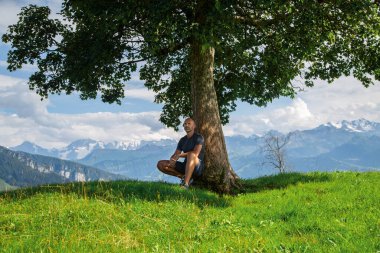 Bir yürüyüşçü tepedeki yeşil bir ağacın altında oturuyor. Arkasında Alpen dağları var.