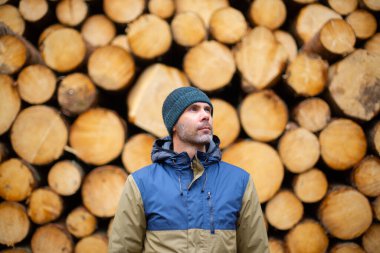 Şapkalı ve ceketli orta yaşlı beyaz adam kütüklerin önünde duruyor.