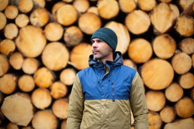 Şapkalı ve ceketli orta yaşlı beyaz adam kütüklerin önünde duruyor.