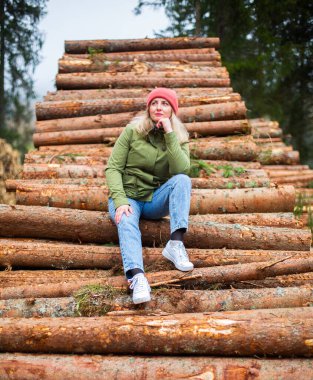 Şapka ve ceket giyen orta yaşlı beyaz kadın ormandaki kütüklerin üzerinde oturuyor.