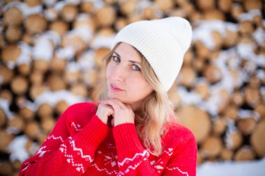 Ormanda kesilmiş odunların önünde geyik ve beyaz şapkalı kırmızı kazaklı mutlu orta yaşlı beyaz kadın. Noel konsepti