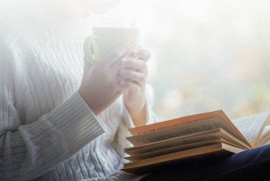 Kız elinde bir fincan çay tutuyor ve pencerenin yanında kitap okuyor.