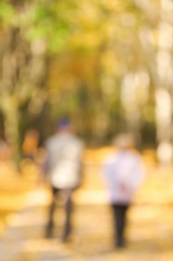 Defocused image of an autumn park with trees, foliage and paths with fallen leaves and people.