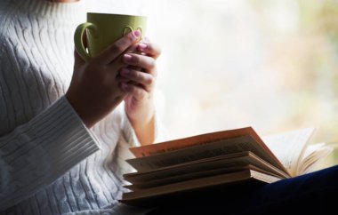 Kız elinde bir fincan çay tutuyor ve pencerenin yanında kitap okuyor.