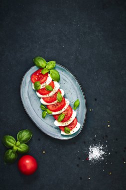 Karanlık masadaki oval tabakta taze salata. Domates, mozzarella, fesleğen ve baharat. Yiyecek ve sağlıklı beslenme konsepti. Yüksek açı görünümü.