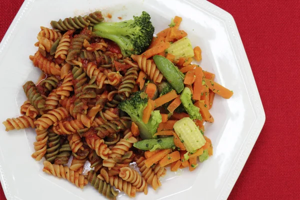 Tri-color rotini pasta with tomato sauce next to vegetable mix of broccoli, corn, baby cob corn, sugar snap peas and carrot. White octagon plate with prepared pasta with tomato sauce and vegetables.