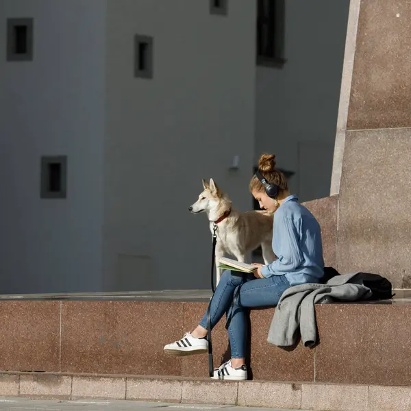 stock image Vilnius, Lithuania - April 22, 2023: A young woman enjoys spending her free time in Cathedral Square near the Gediminas Monument. She is reading a book and listening to music. Dog is standing nearby.