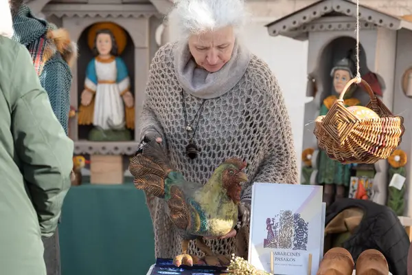 Vilnius, Litvanya - 8 Mart 2025: Litvanya 'nın Vilnius kentindeki Kaziukas fuarında el yapımı ahşap horoz ve halk sanatı satan yaşlı zanaatkar.