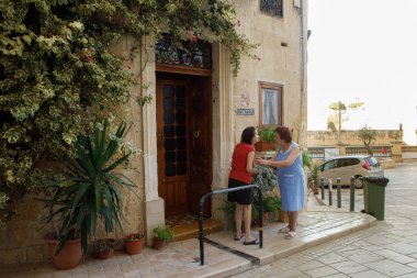 Birgu, Malta - 13 Ekim 2023: İki yaşlı kadın Ave Maria Caddesi 'ndeki geleneksel kireçtaşı bir evin önünde sıcak ve hareketli bir sohbete girişiyor. Ev bereketli saksı bitkileriyle süslenmiş. Bakire Meryem 'in dini bir plaketi..