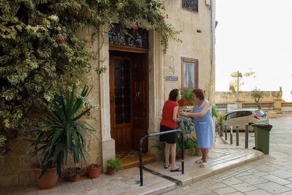 Birgu, Malta - 13 Ekim 2023: İki yaşlı kadın Ave Maria Caddesi 'ndeki geleneksel kireçtaşı bir evin önünde sıcak ve hareketli bir sohbete girişiyor. Ev bereketli saksı bitkileriyle süslenmiş. Bakire Meryem 'in dini bir plaketi..