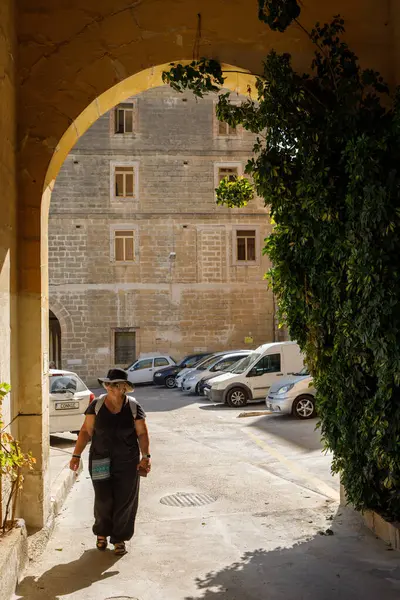 Senglea, Malta - 13 Ekim 2023: Geleneksel kireçtaşı mimarisi ve park halindeki araçlarla çevrili Senglea 'da güneşli bir taş kemer yolunda yürüyen günlük kıyafetli bir kadın.