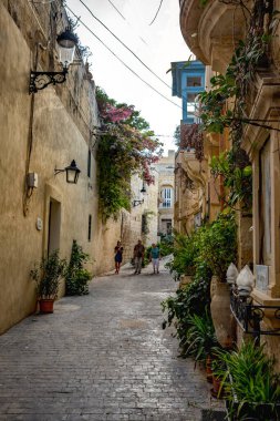 Rabat, Malta - 8 Ekim 2023: Ziyaretçiler tarihi kireçtaşı binalar, renkli balkonlar, asma tesisleri ve geleneksel mimariyle çevrili sakin, kaldırımlı bir sokak boyunca yürüyorlar..