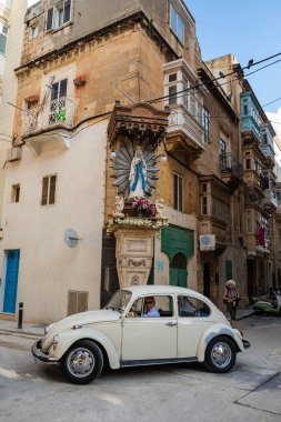 Floriana, Malta - 8 Ekim 2023: Klasik beyaz bir Volkswagen Beetle, Floriana 'da Valletta' nın bitişiğindeki bir banliyöde bulunan geleneksel bir Madonna tapınağının yanından geçiyor.. 