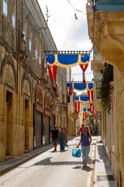 Rabat, Malta - 8 Ekim 2023: Yerel halk ve ziyaretçiler, mavi, kırmızı ve altın renkli canlı festa afişlerle süslenmiş Malta 'nın Rabat kentinde dar bir sokakta yürüyorlar.. 