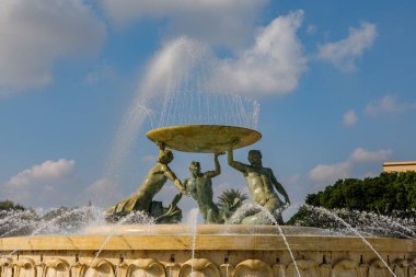 Valletta, Malta 'daki Triton Çeşmesi, üç bronz figürle büyük bir leğen tutuyor, parlak mavi gökyüzünün altında su püskürtüleriyle çevrili..