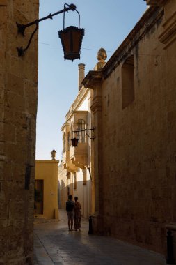 Mdina, Malta - 8 Ekim 2023: Güneş ışığı Mdina 'da tarihi ve dar bir ara sokağı aydınlatarak antik taş duvarları ve geleneksel bir tepedeki feneri aydınlatıyor..