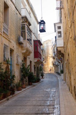 Birgu, Malta - 13 Ekim 2023: Geleneksel Malta balkonları ve asılı bir fenerin yer aldığı tarihi binaların arasından büyüleyici dar bir kaldırım taşı caddesi esiyor.