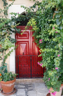 Karmaşık bir demir ızgara ile korunan parlak kırmızı bir Malta kapısı Senglea, Malta 'daki yemyeşil bougainvillea bitkileri tarafından çerçevelenmiştir.