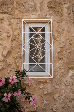  Bir dekoratif beyaz güvenlik ızgarası, Rabat, Malta 'daki canlı pembe amber çiçekleri ile tamamlanmış geleneksel bir taş binanın penceresini süslüyor.