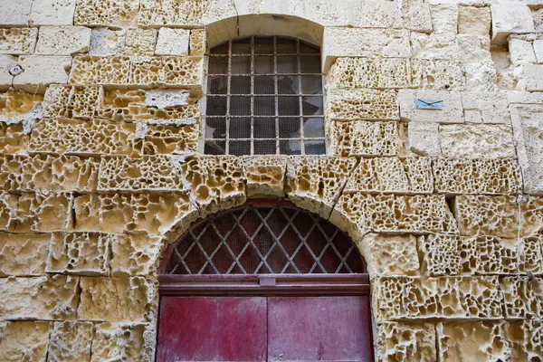 Kalıplı, gözenekli taş bir duvar, Birgu, Malta 'da kavisli, elmas desenli ızgaralı kırmızı bir kapının üzerinde rendelenmiş bir pencere içerir.