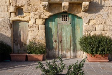Marsaxlokk, Malta 'da saksıda bitkiler bulunan eski, dokulu taş bir duvara yerleştirilmiş iki kırsal, yıpranmış kapı - biri yeşil ve biri doğal ahşap