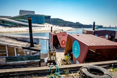 Oslo, Norveç - 18 Temmuz 2025: Geleneksel tasarımların yer aldığı eşsiz yüzen saunalar ve hamamlar, Oslo 'da canlı bir rıhtım boyunca demirlemiş ve arka planda modern mimariyle benzersiz bir eğlence deneyimi sunmaktadır..