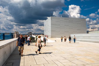 Oslo, Norveç - 15 Temmuz 2025: Ziyaretçiler Oslo Opera Binası 'nın eşsiz eğimli çatısını keşfederek, parçalı bulutlu bir gökyüzü altında modern mimari ve panoramik şehir manzaralarının keyfini çıkarıyorlar..