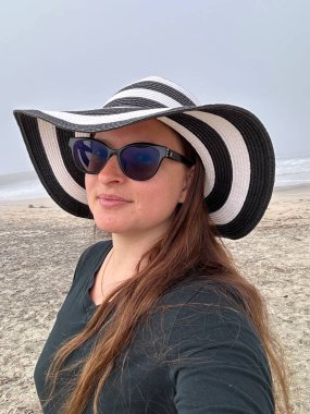 Huzurlu bir fotoğraf, siyah beyaz bir plaj şapkası ve koyu renk bir tişörtle okyanusun kenarında duran bir kadını yakalar..