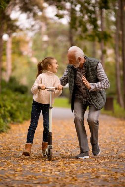 Son sınıf öğrencisi, torununa sonbahar parkında scooter sürmeyi öğretiyor.