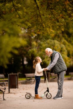 Son sınıf öğrencisi, torununa sonbahar parkında scooter sürmeyi öğretiyor.