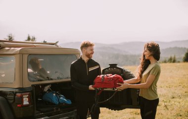 Smiling young couple preparing hiking adventure with backpacks by terrain vehicle