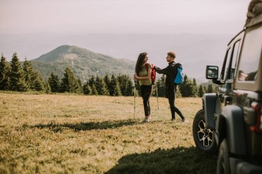 Smiling young couple preparing hiking adventure with backpacks by terrain vehicle