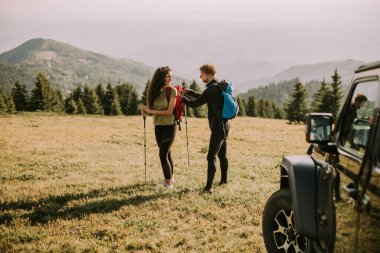 Smiling young couple preparing hiking adventure with backpacks by terrain vehicle