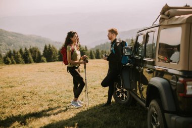 Smiling young couple preparing hiking adventure with backpacks by terrain vehicle