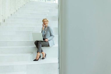 İş kadını, modern ofis merdivenlerinde dizüstü bilgisayarla çalışırken cep telefonu kullanıyor.