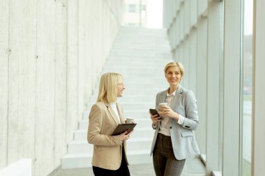 Ofis koridorunda dijital tablet ve cep telefonuyla yürüyen iki iş kadını.